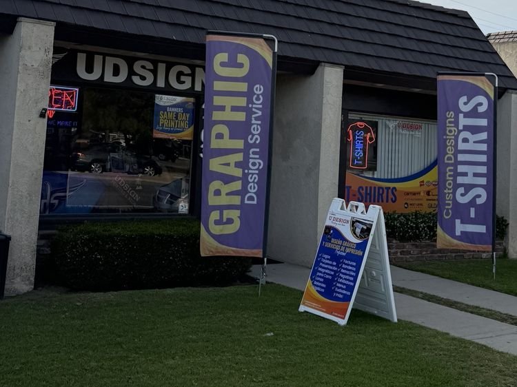 The U Design storefront on a low tile-roofed building, with "Graphic Design Service" and "Custom Designs T-Shirts" flags and an A-frame sign on the lawn.