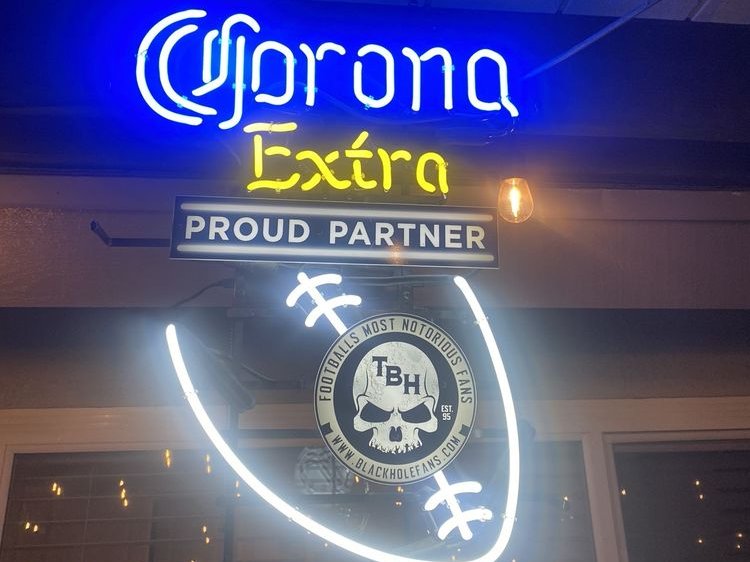 A lit blue and yellow beer neon above a white football-shaped neon with a skull badge, mounted under a porch roof at night.
