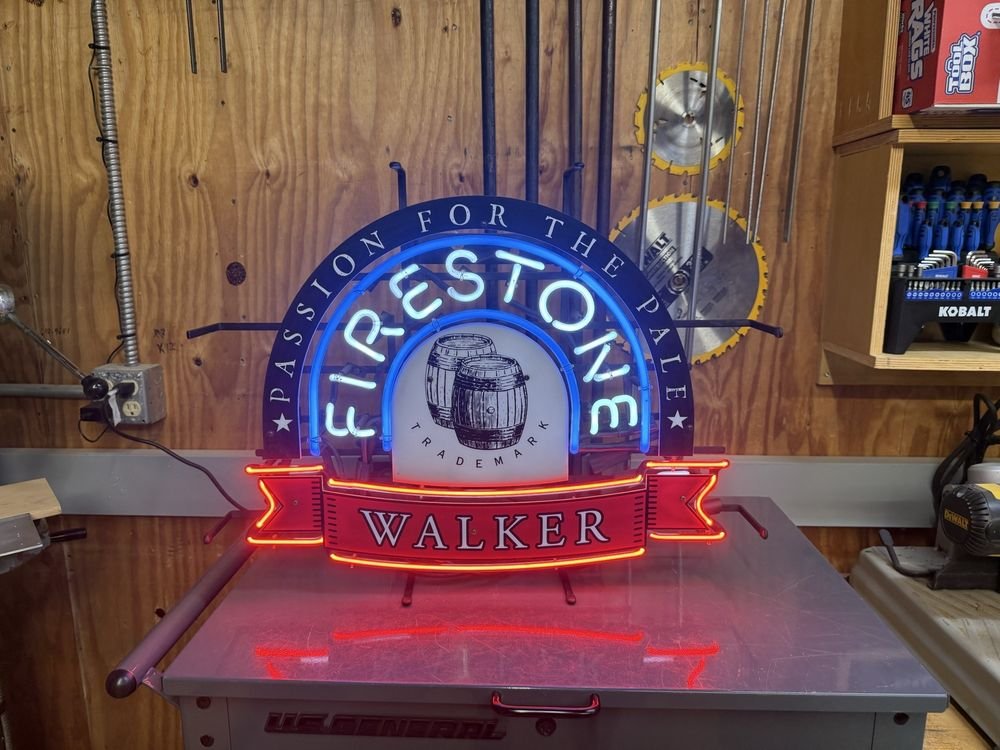 A lit neon sign with a blue arch of lettering and a red ribbon panel, standing on a metal tool chest against a plywood wall.