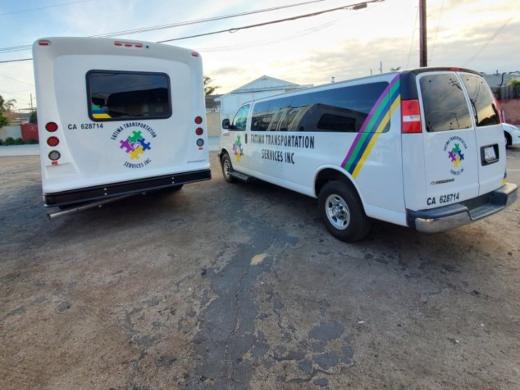 A white passenger van with purple, green, blue and yellow stripes and a puzzle-piece logo, parked beside a matching shuttle bus.