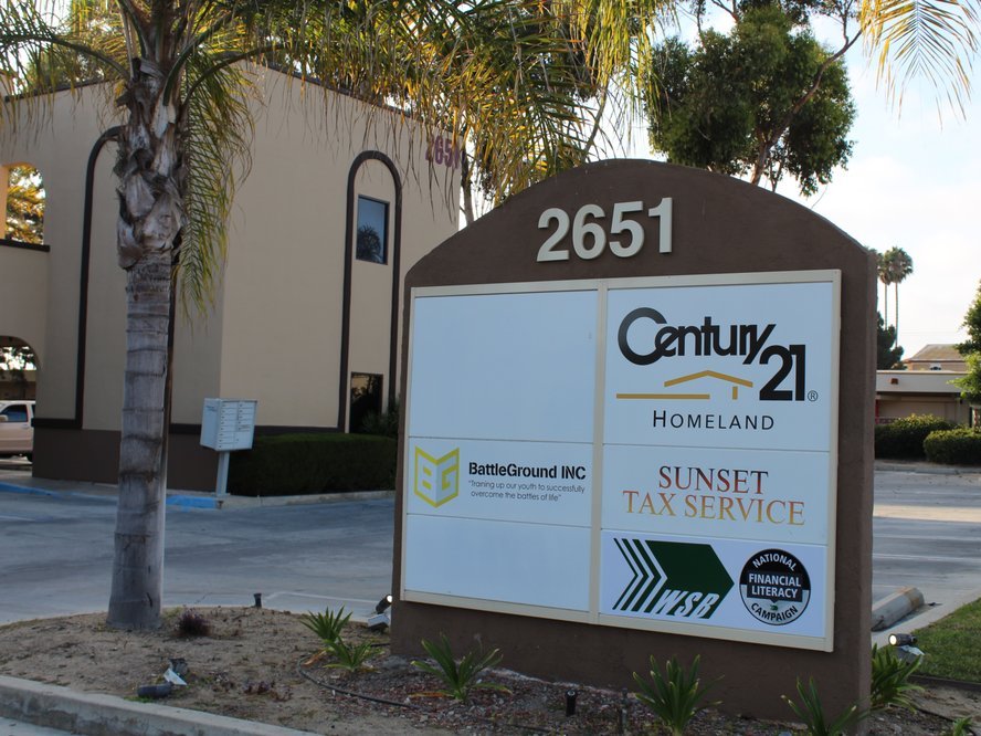 A brown monument sign with the address number on top and printed tenant panels, in front of an office building with palms.