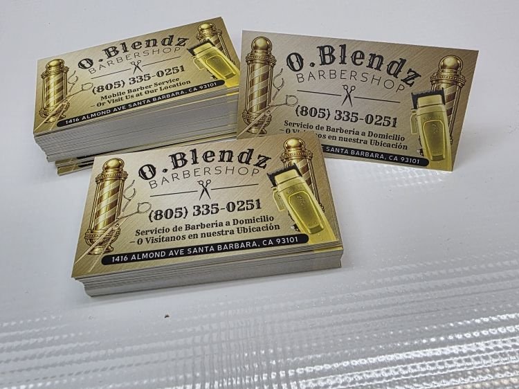 Stacks of business cards printed with a gold barber-pole and clipper design, a phone number and an address in English and Spanish.